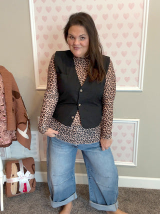 Woman wearing a leopard print top and black vest with rolled-up jeans in a room with heart-patterned wallpaper.