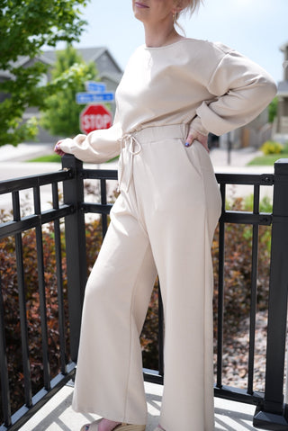 Person wearing a beige one shoulder jumpsuit standing on a balcony with a blurred background
