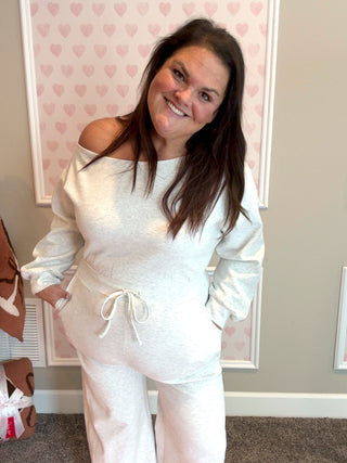 Woman wearing a ash gray one shoulder jumpsuit outfit in a room with heart-patterned wallpaper.