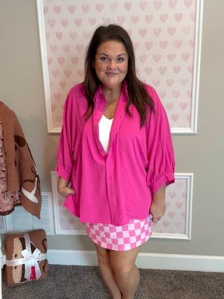 Woman wearing a bright pink shirt and checkered skirt in a room with heart-patterned wallpaper.