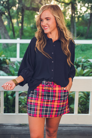 Woman wearing a navy shirt and plaid skirt standing outdoors with greenery in the background