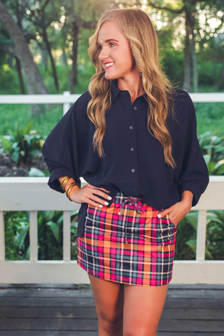 Woman wearing a black shirt and plaid skirt standing outdoors with greenery in the background