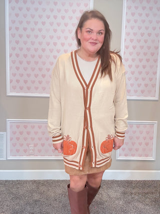Woman wearing a beige cardigan with pumpkin pockets in a room with heart-patterned wallpaper.