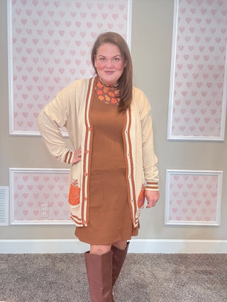 Woman wearing a brown dress with a beige cardigan in a room with heart-patterned wallpaper.