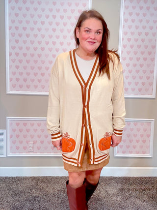 Woman wearing a beige cardigan over a dress with pumpkin pockets, standing in front of a wall with heart patterns.