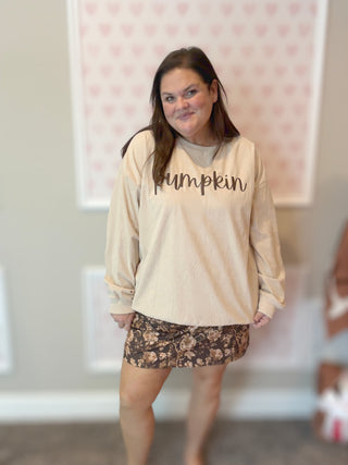 Woman wearing a beige sweatshirt with 'pumpkin' text and a floral skirt indoors.