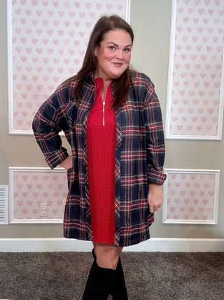Woman wearing a red dress and plaid flannel in front of a wall with heart patterns.