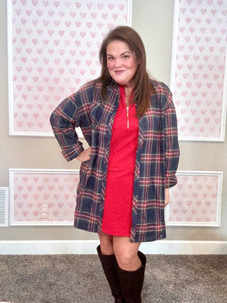 Woman wearing a plaid flannel over a red dress with knee-high boots in a room with heart-patterned wallpaper.