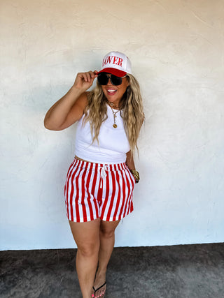 Model walking outdoors in striped shorts, holding sunglasses for a casual summer vibe.