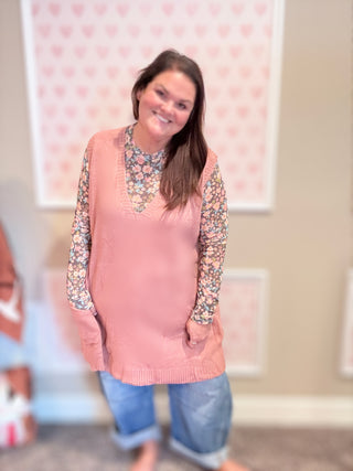 Woman wearing a pink sweater vest over a floral long-sleeve shirt with blue jeans, standing in a room with a heart-patterned wall.