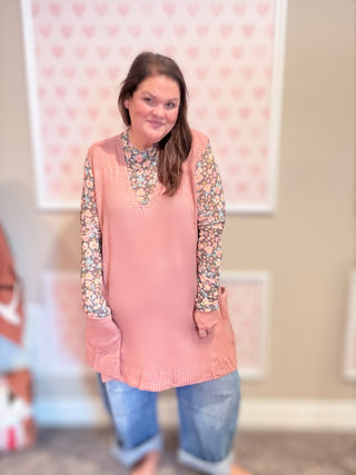 Woman wearing a pink sweater with floral sleeves in a room with a heart-patterned curtain.