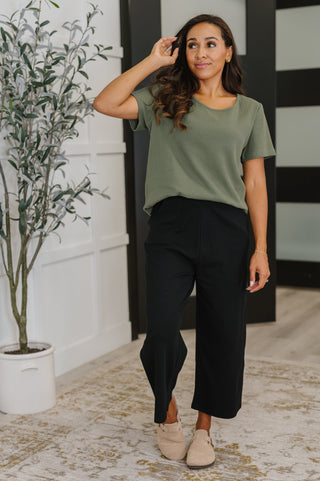 Woman wearing a green top and black pants standing indoors with a plant in the background