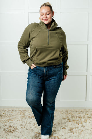Person wearing an olive green pullover and blue jeans against a white paneled wall.