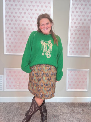 Person wearing a green sweater with a horse design and a western patterned skirt, standing in front of a wall with heart designs.