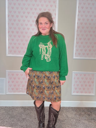 Person wearing a green sweater with a horse design and a western patterned skirt, standing in a room with heart-shaped wallpaper.