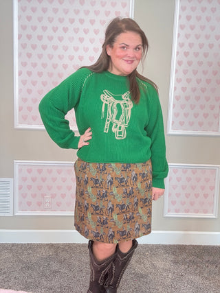 Woman wearing a green sweater with a graphic design and a western patterned skirt, standing in a room with heart-shaped decorations on the wall.