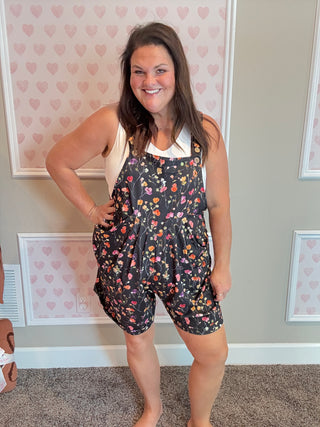 Woman wearing a floral overalls standing in a room with heart-patterned wallpaper.