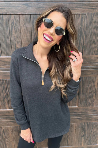 Woman wearing a faded black zip-up sweatshirt and sunglasses against a wooden wall.