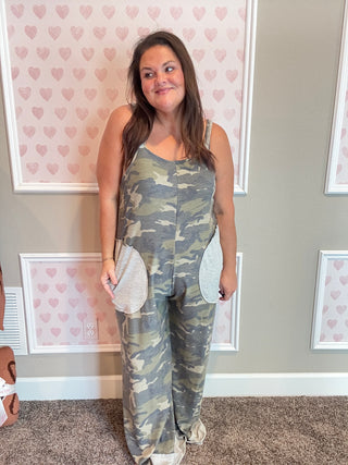 Woman wearing a camouflage jumpsuit standing in a room with heart-patterned wallpaper.
