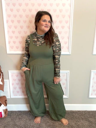 Woman wearing a green jumpsuit with camouflage sleeves in a room with heart-patterned wallpaper.