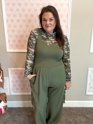 Woman wearing a green jumpsuit with camouflage sleeves in a room with heart-patterned wallpaper.