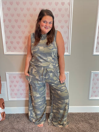 Woman wearing a camouflage jumpsuit standing in front of a wall with heart patterns.