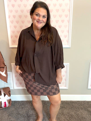 Woman wearing a brown shirt and checkered skirt standing in front of a wall with heart patterns.