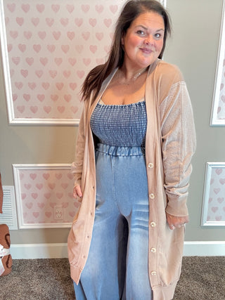 Woman wearing a beige cardigan over a blue jumpsuit in a room with heart-patterned wallpaper.