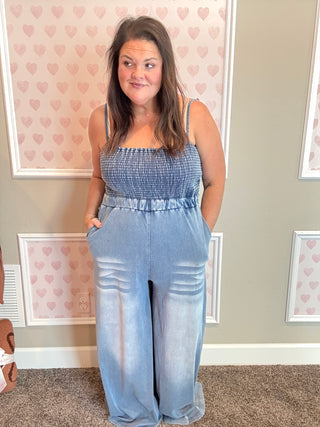 Woman wearing a blue jumpsuit standing in a room with heart-patterned wallpaper.