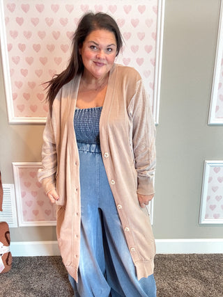 Woman wearing a long beige cardigan over a blue dress in a room with heart-patterned wallpaper.