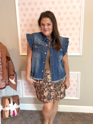 Woman wearing a denim vest over a floral skirt in a room with heart-patterned wallpaper.