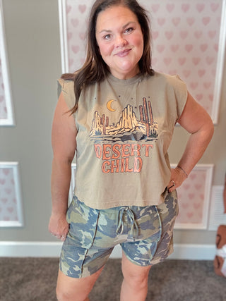 Woman wearing a 'Desert Child' graphic t-shirt and camouflage shorts in a room with heart-patterned wallpaper.