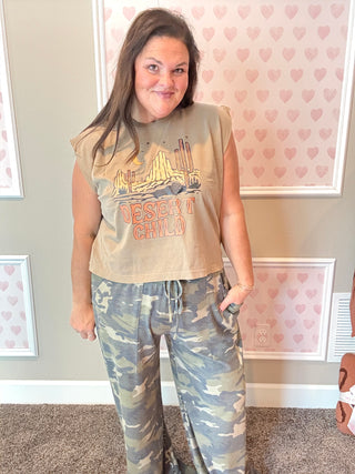 Woman wearing a sleeveless graphic tee and camouflage pants in a room with heart-patterned wallpaper.