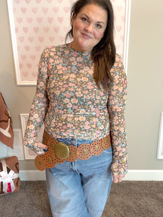 Woman wearing a floral long-sleeve mesh top and jeans with a belt in a room with heart-patterned wallpaper.