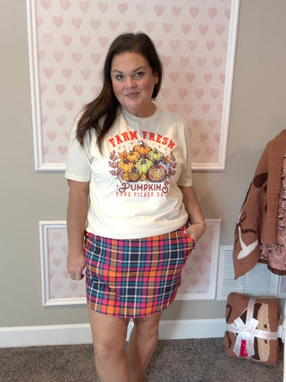Woman wearing a 'Farm Fresh Pumpkins' t-shirt and plaid skirt in a room with heart-patterned wallpaper.