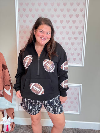 Woman wearing a black hoodie with football designs and patterned shorts in front of heart-patterned wallpaper.