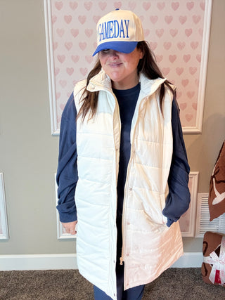 Person wearing a white puffer vest over a navy long-sleeve shirt, with a 'GAMEDAY' cap, in a room with heart-patterned wallpaper.