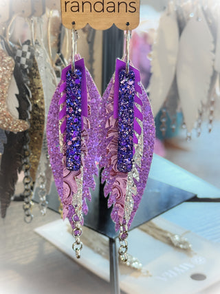 Layered dangle earrings in rich purple and lavender tones with sparkling glitter, paisley patterns, and silver chain accents. Hanging from a “randans” card on a rotating earring display.