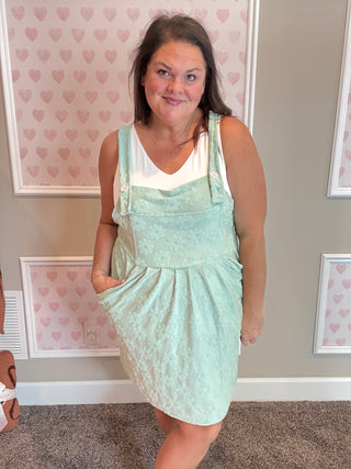 Woman wearing a light green dress with a white top in a room with heart-patterned wallpaper.
