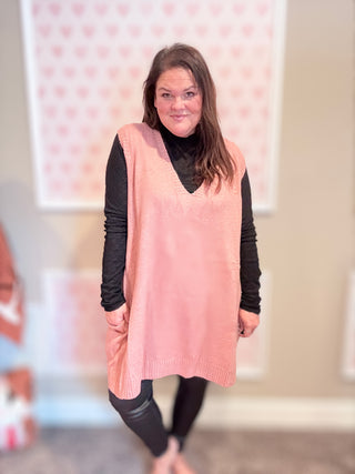 Woman wearing a pink sweater vest over a black long-sleeve shirt and leggings indoors.