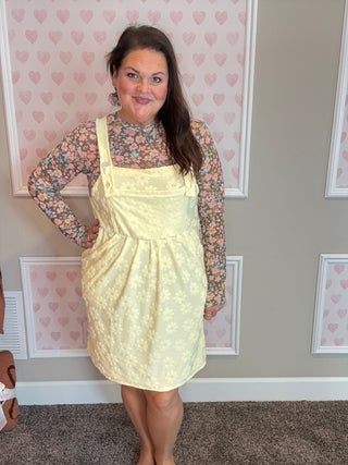 Woman wearing a yellow pinafore dress over a floral top in a room with heart-patterned wallpaper.