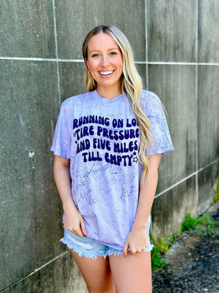 Woman wearing a tie-dye t-shirt with text standing against a wall.