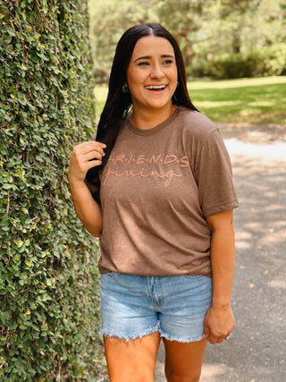 Woman wearing a brown t-shirt with 'FRIENDS' printed on it, standing outdoors next to a hedge.