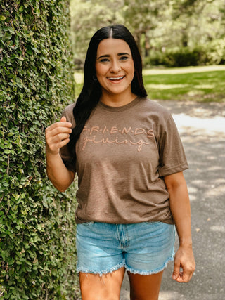 Woman wearing a brown t-shirt with text, standing next to a green hedge.