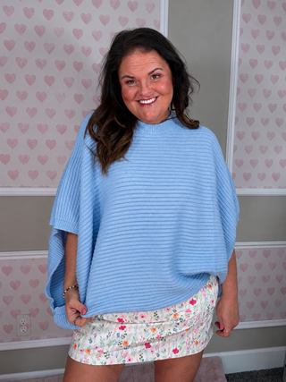 A woman wearing a light blue ribbed knit poncho sweater and a white floral mini skirt, standing in front of a wall with pink heart wallpaper.