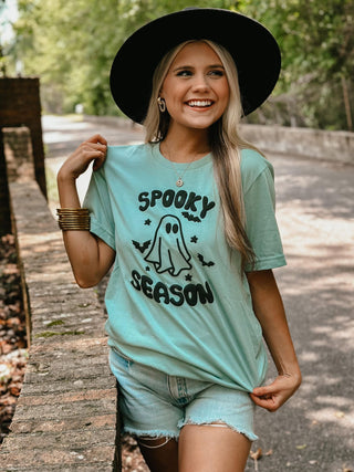 Woman wearing a light blue t-shirt with 'Spooky Season' graphic and black hat outdoors.