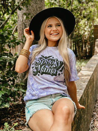 Woman wearing a tie-dye t-shirt with 'Spooky Mama' text, sitting outdoors.