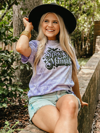 Woman wearing a 'Schoo Mama' t-shirt sitting outdoors among trees.
