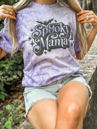 Person wearing a purple tie-dye t-shirt with 'Spooky Mama' text, sitting outdoors.
