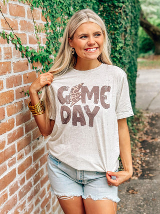 Woman wearing a 'Game Day' t-shirt standing against a brick wall with greenery.
